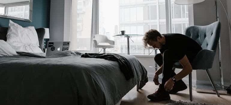 A person kneels on a bedroom floor, tying their shoes near a neatly made bed and large windows overlooking a cityscape.